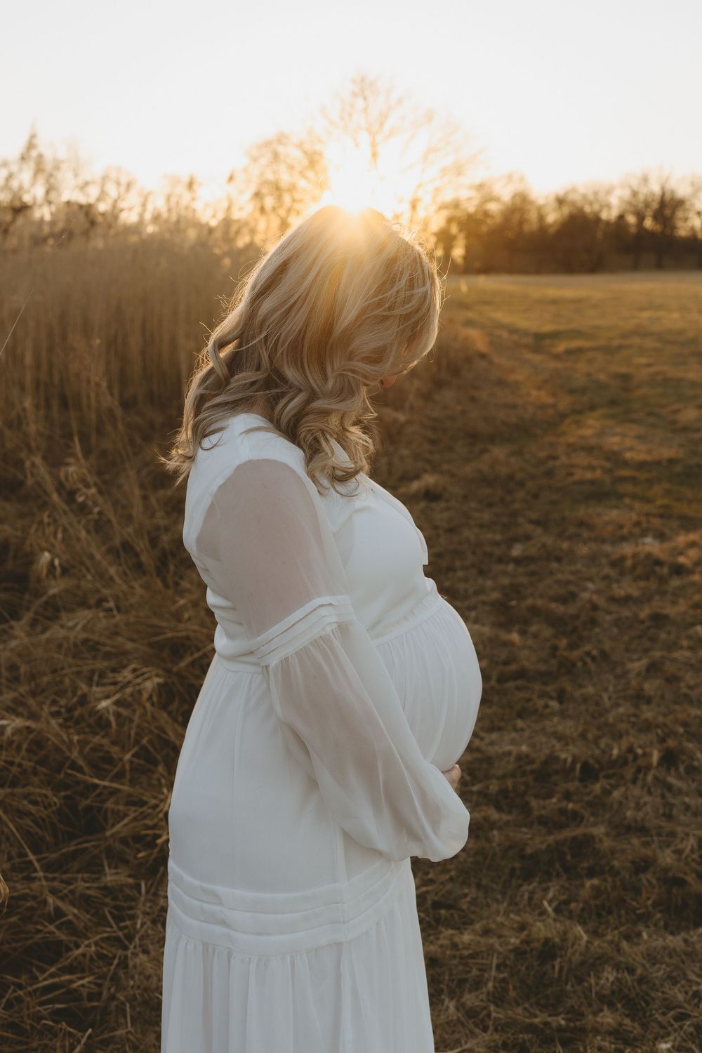 Pregnant woman in flowing white dress stands in golden sunlit field during sunset maternity photoshoot.