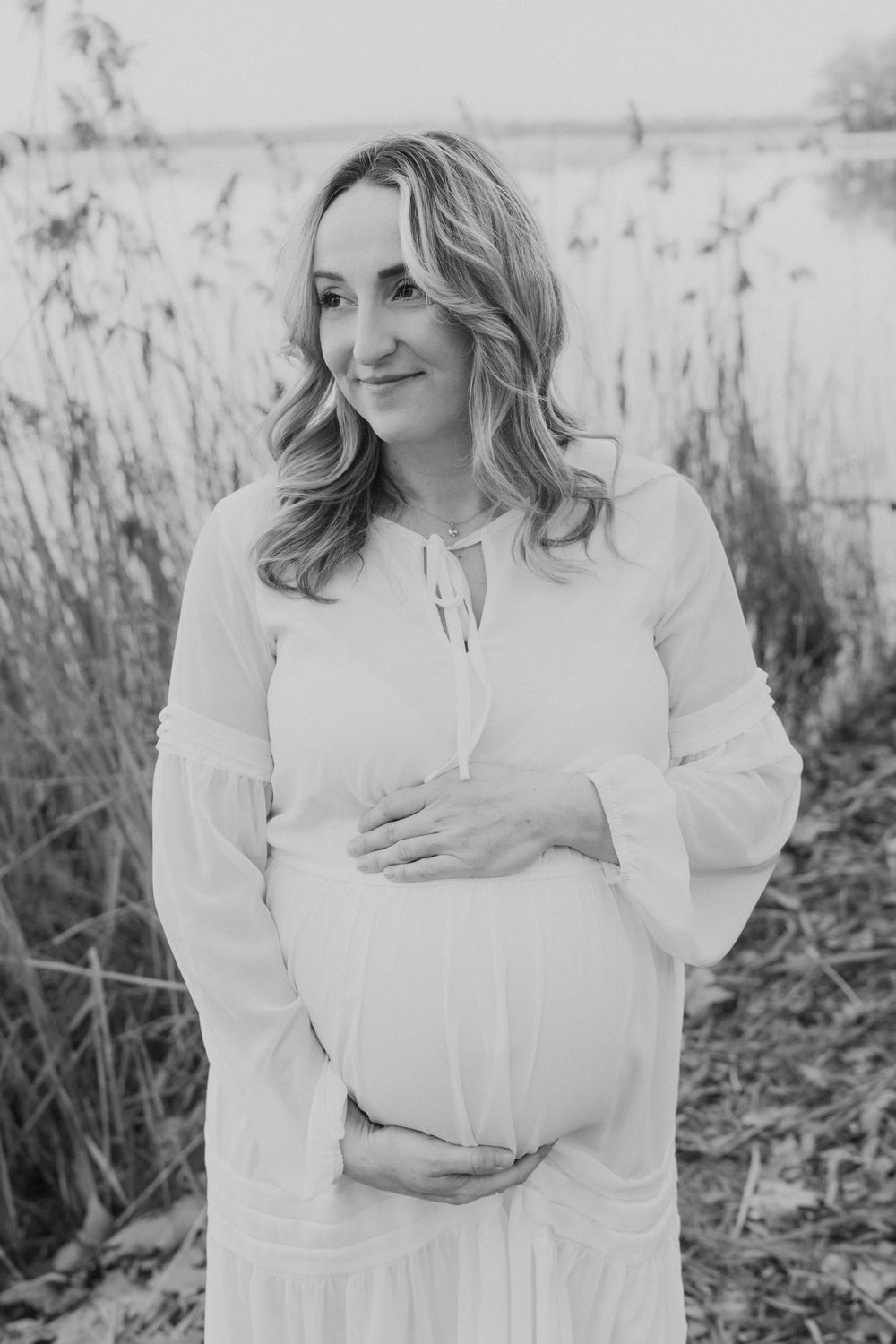 Expectant mother in a flowing white dress poses in front of tall grass in black and white photography.