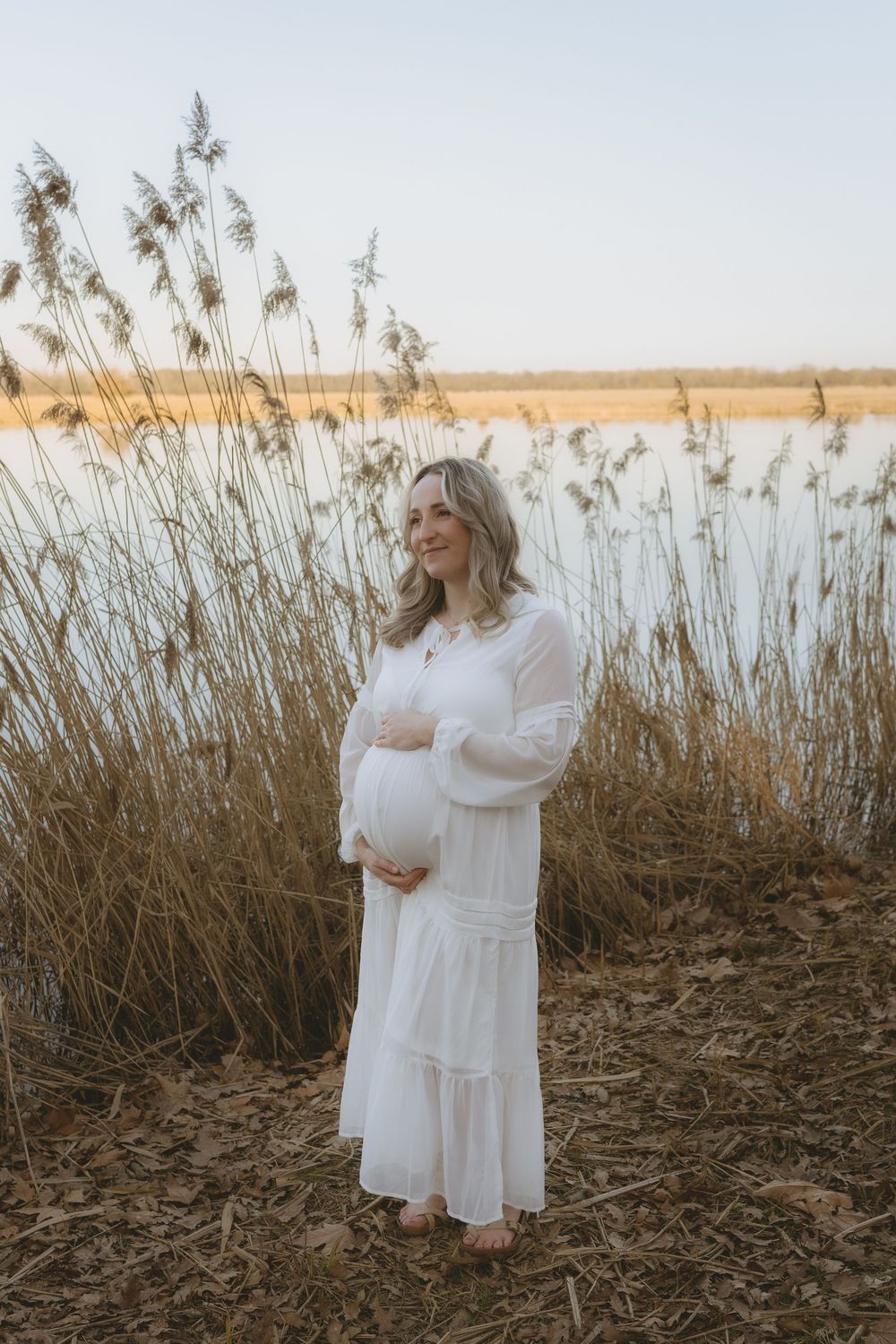 Ethereal maternity scene with expecting mother in white dress standing among tall reeds at golden hour by the water.