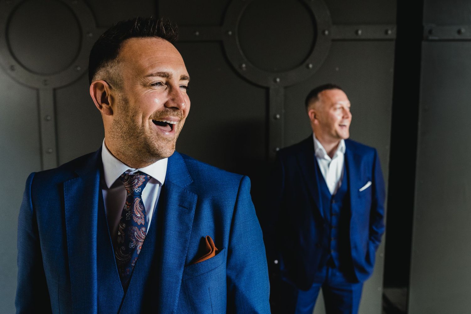 Two men wearing blue suits smile while posing against a gray wall in an elegant setting.