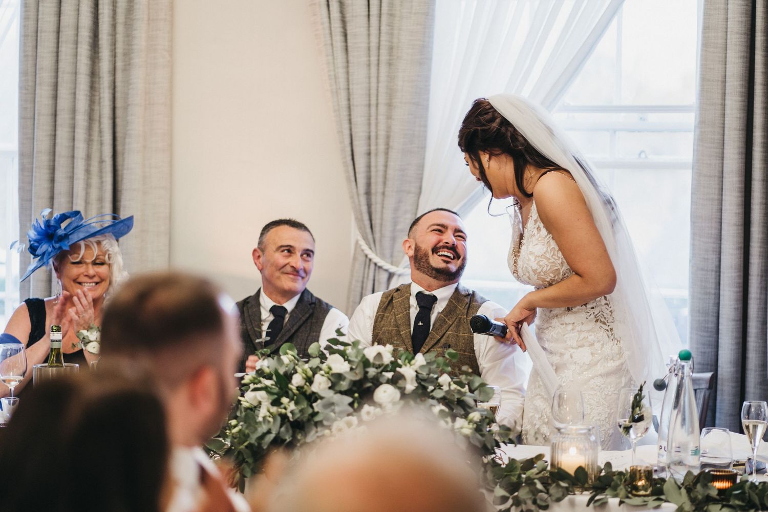 Two people share a joyful moment during their wedding reception at a table decorated with white floral arrangements.