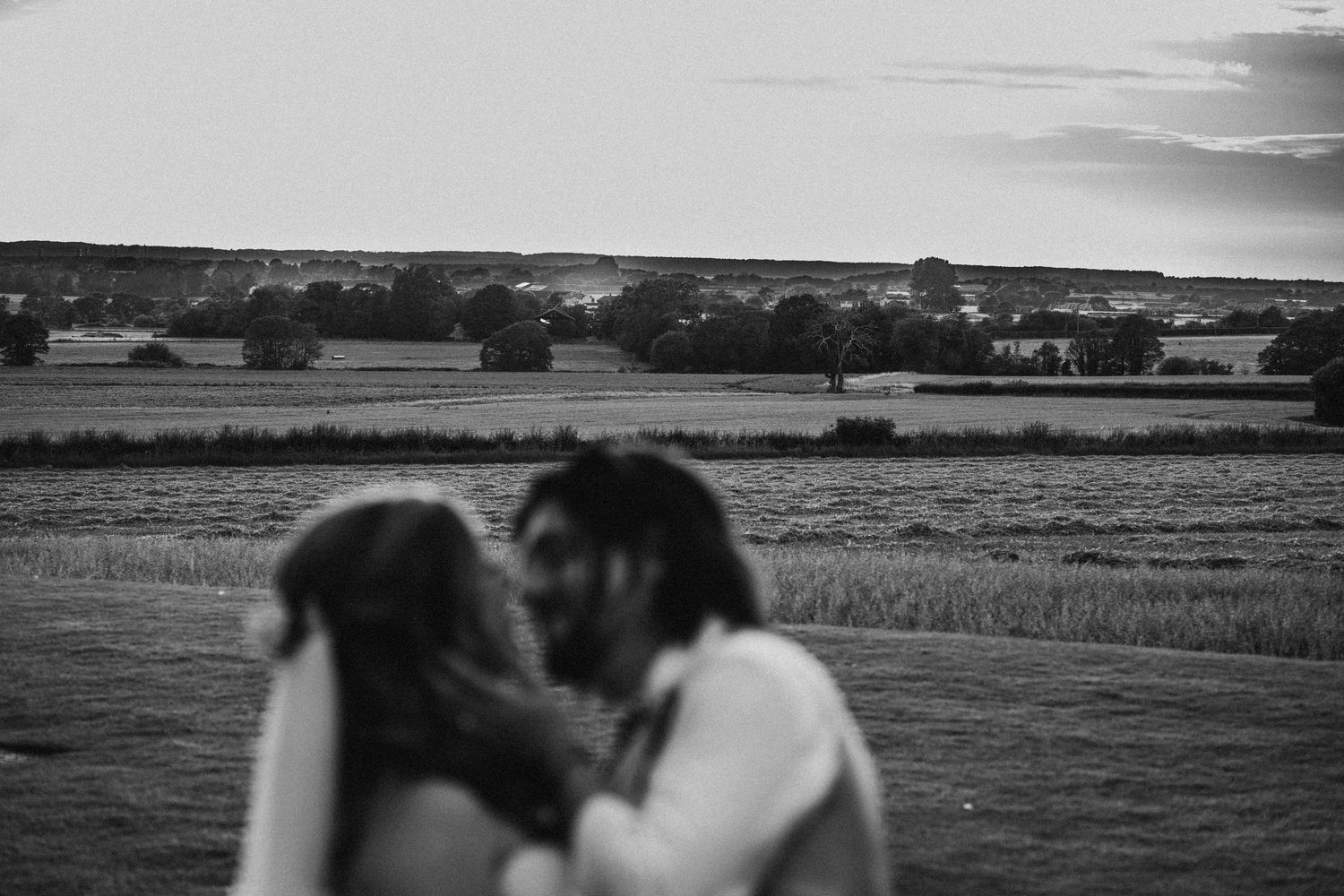 A romantic couple embraces in black and white against a rural countryside landscape at dusk.