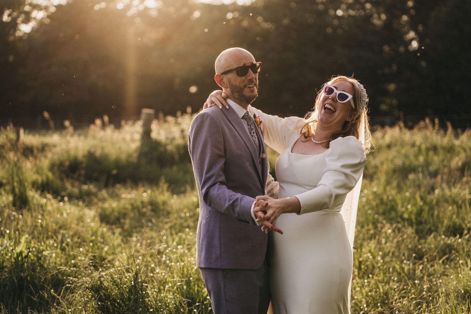 Happy couple sharing laughter together in a sunlit meadow during golden hour wearing sunglasses and formal attire.