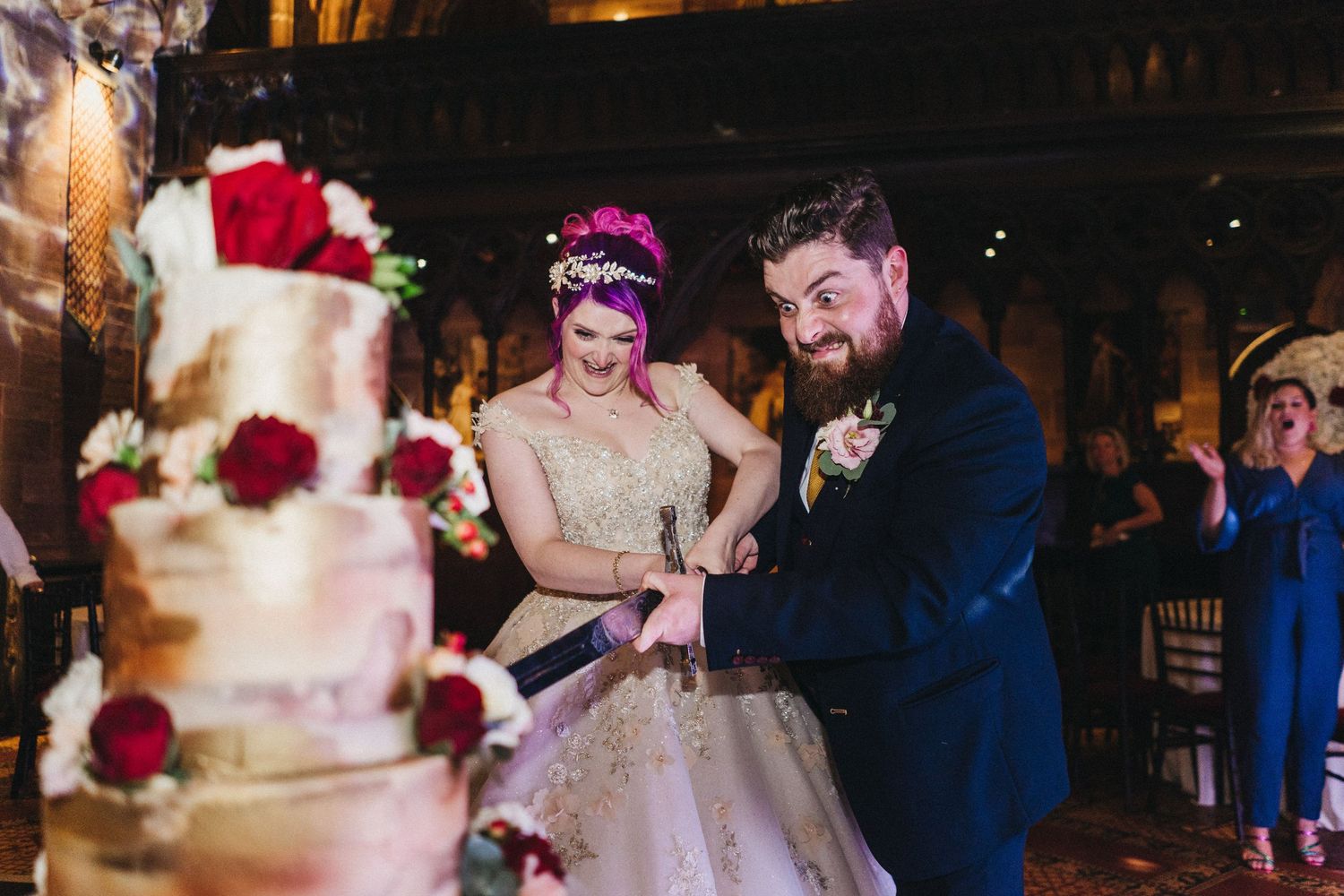 Joyful couple cutting their elegant multi-tiered wedding cake with red roses at their evening reception celebration.