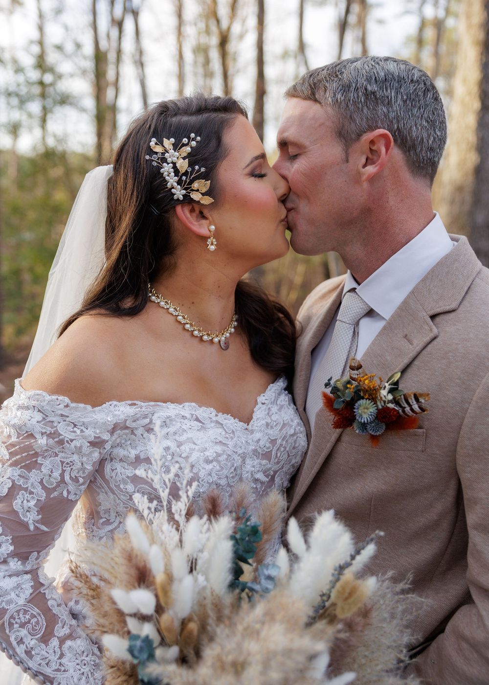 Bride and groom sharing a kiss outdoors, bride wearing lace gown, veil, and floral hair piece, groom in tan suit with boutonniere.