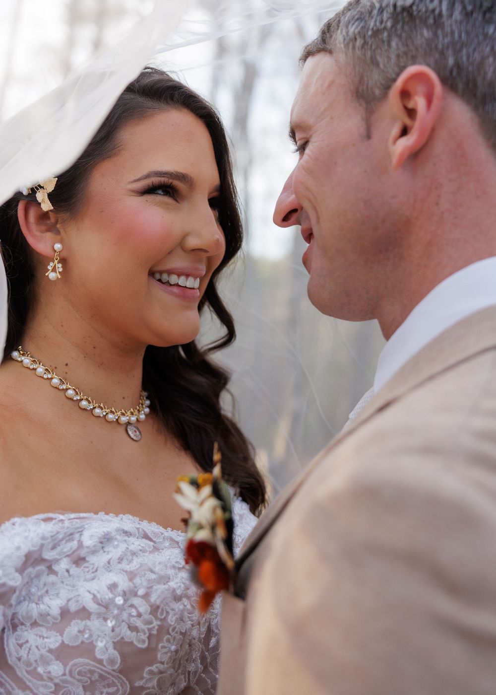 A smiling bride in a lace gown and veil gazes at her groom in a tan suit during an outdoor wedding ceremony.