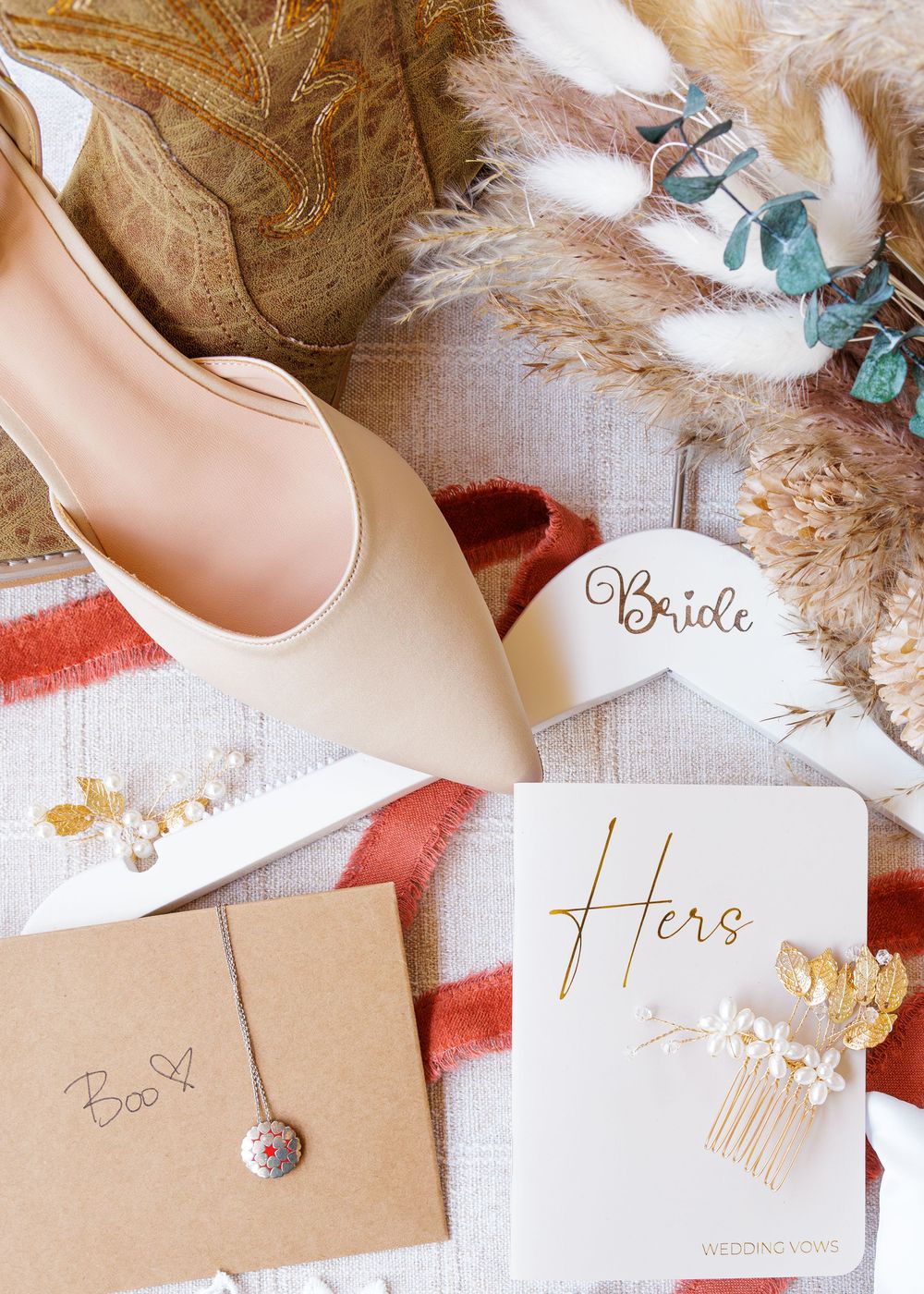 Bridal flat lay featuring nude heels, Bride mug, Hers vow book, floral hair comb, necklace, and dried pampas grass.