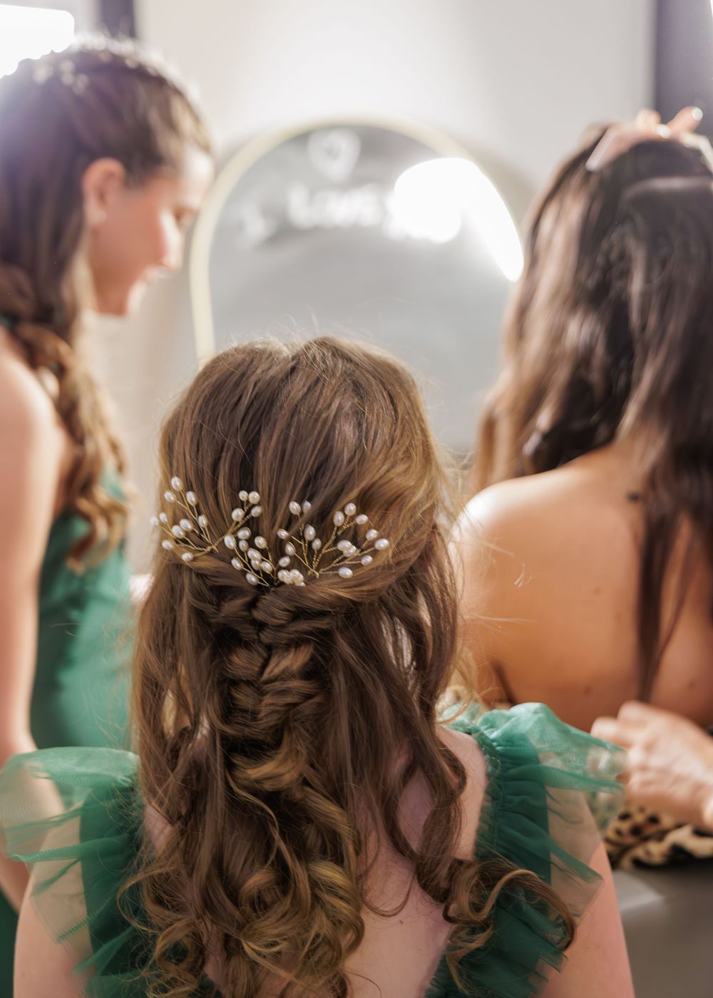 Bridesmaids in green dresses with braided half-up hairstyle adorned with pearl hair pins near a mirror.