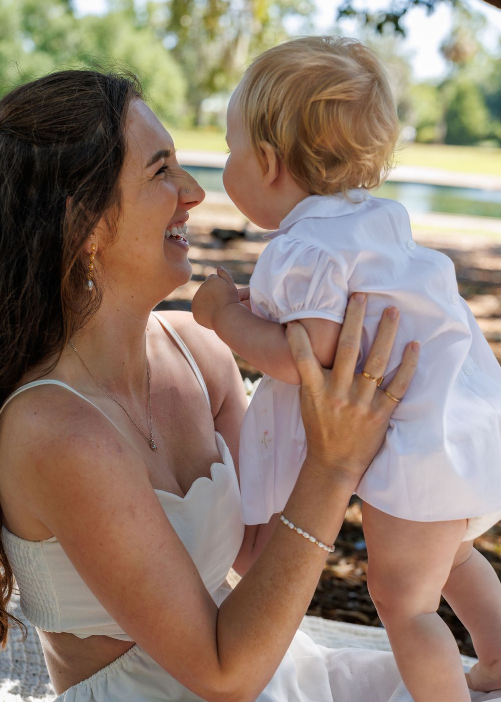 A smiling woman in a white dress holds a baby in a white outfit outdoors in a sunny park setting.