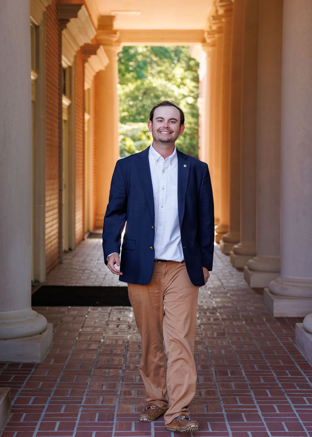 Young man in navy blazer and khaki pants walking confidently through a brick columned hallway outdoors.