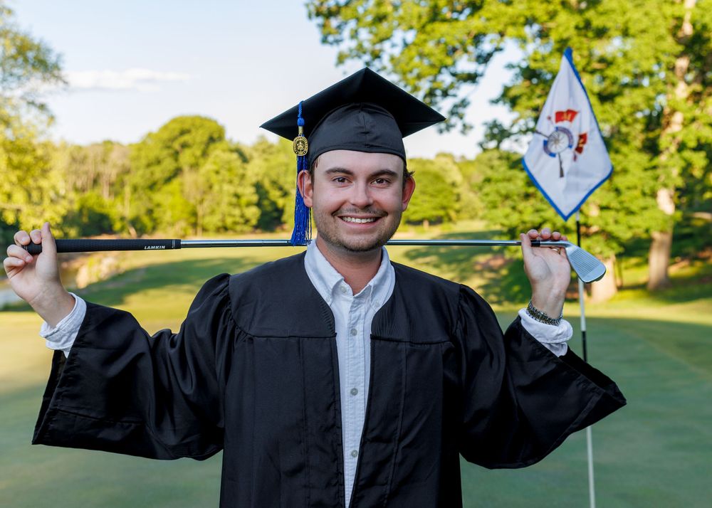 Smiling male graduate in cap and gown holding a golf flag on a green course on a sunny day.