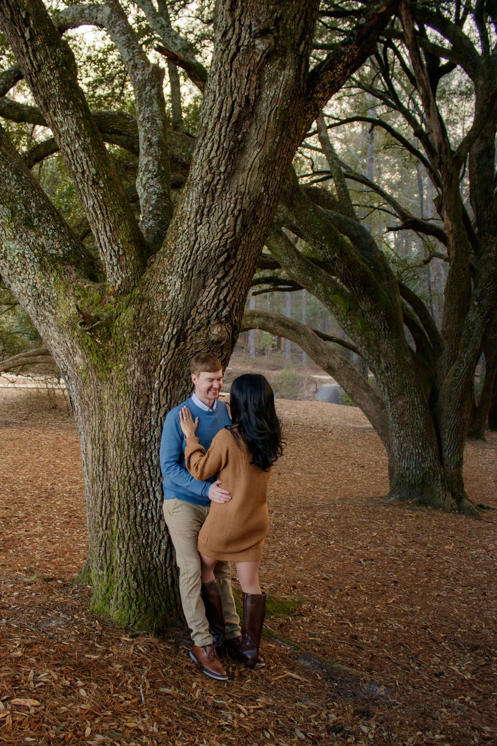 Couple sharing a romantic moment leaning against a large oak tree surrounded by autumn leaves in a forest.