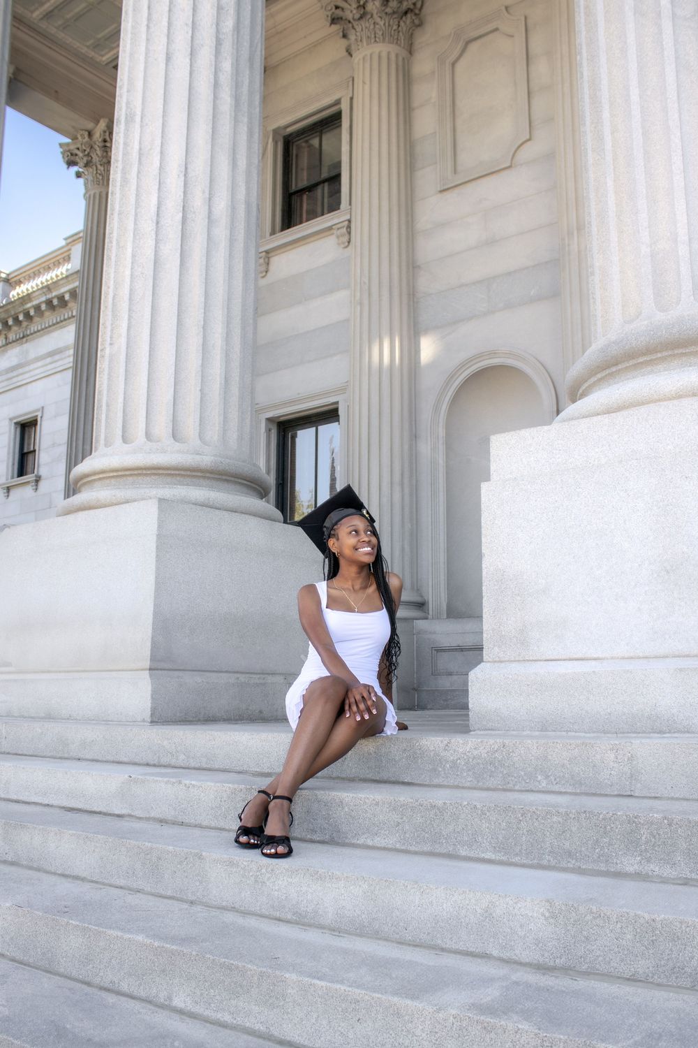 Young woman in white dress and graduation cap sitting on steps of a grand building with tall columns.