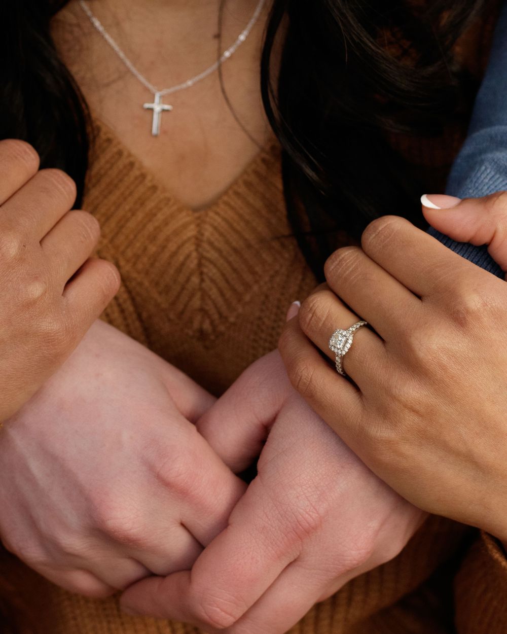 Close-up of hands with engagement ring and cross necklace against brown sweater background.