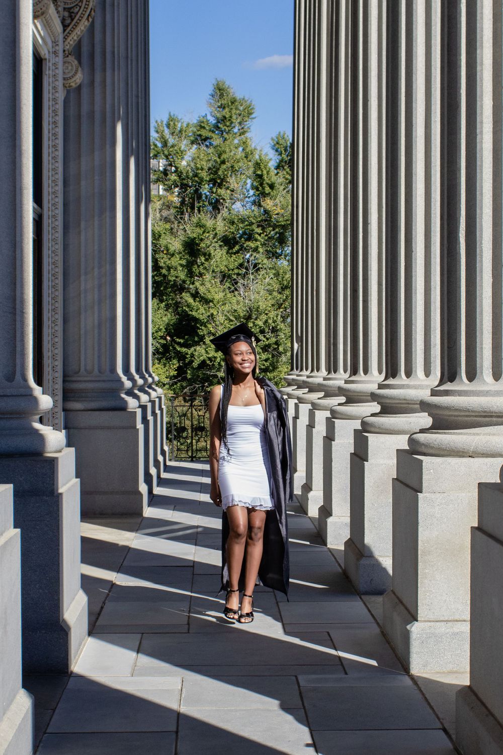 A graduate in white dress poses between classical columns on a sunny day.