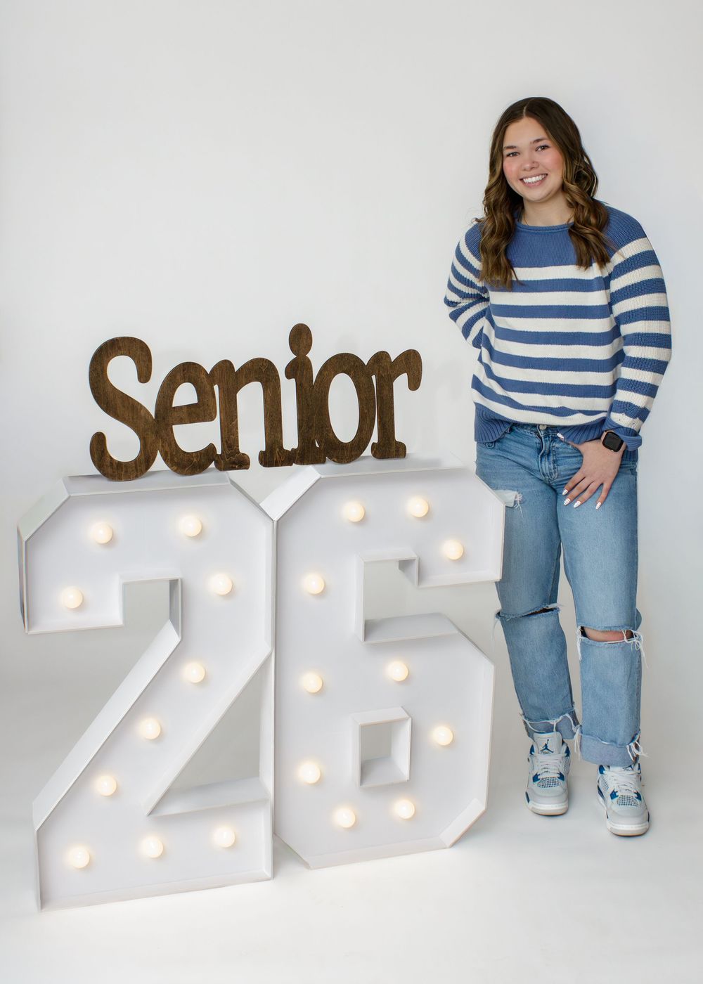 A student in a blue striped sweater and ripped jeans poses with illuminated numbers '25' and 'Senior' text display.