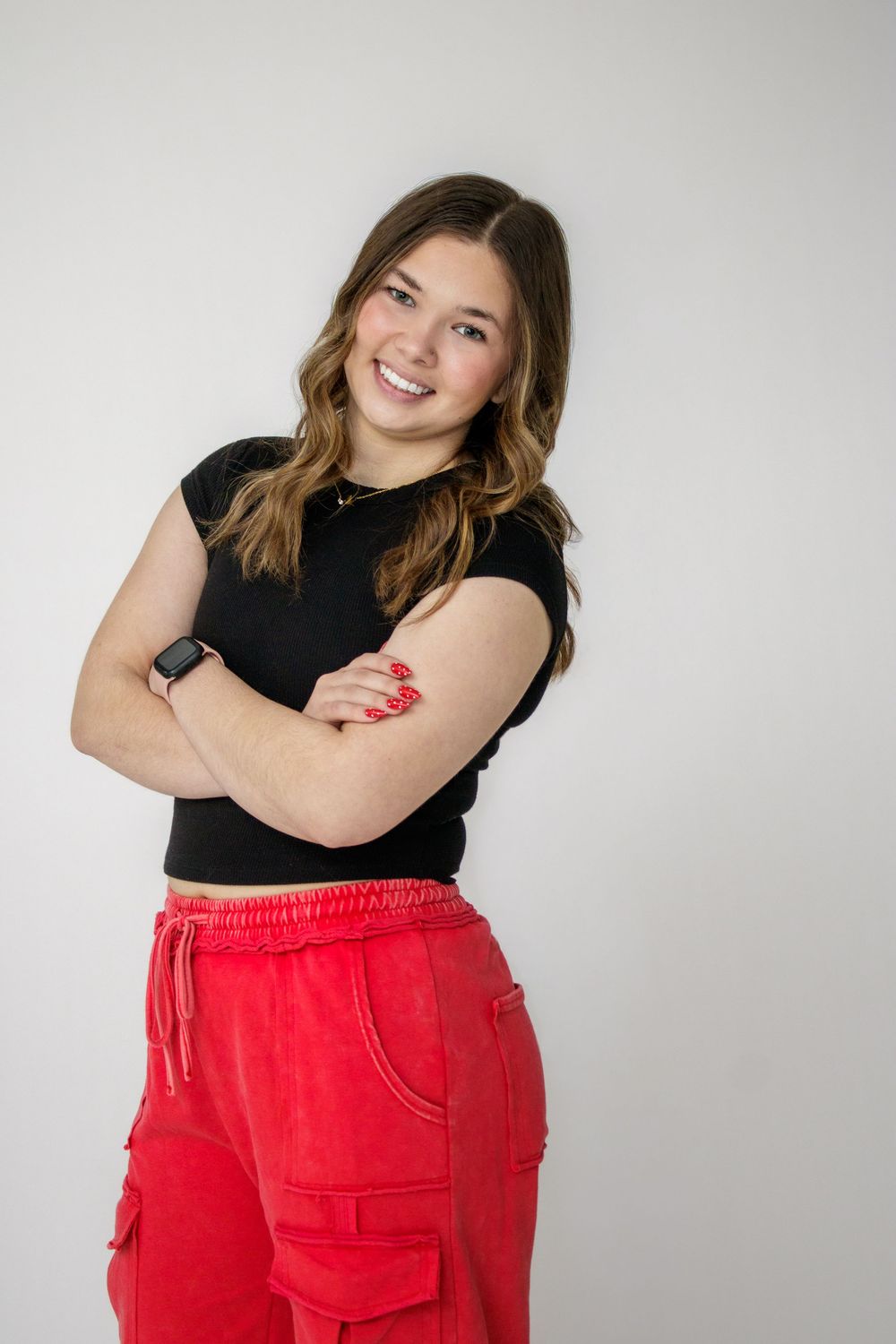 A person wearing a black top and bright red pants poses confidently against a white studio background.