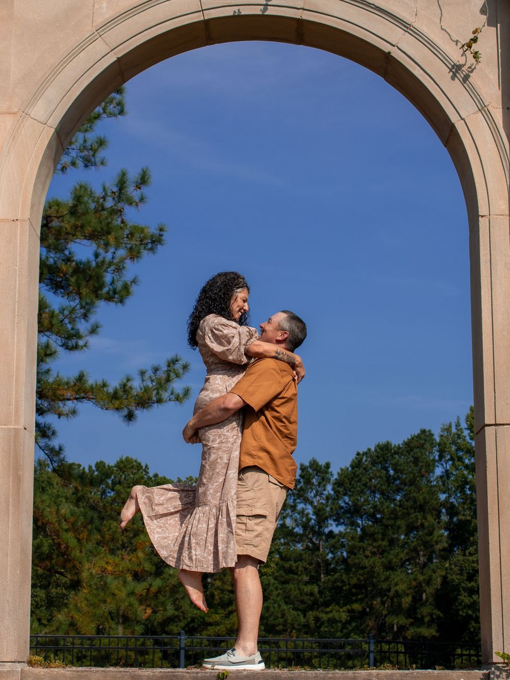 A couple poses playfully under a stone archway on a sunny day.