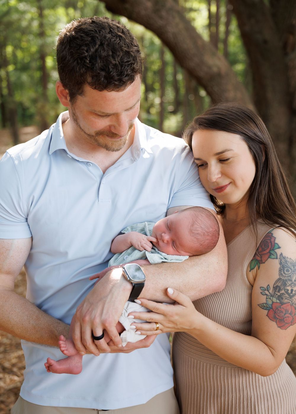 New parents tenderly hold their sleeping newborn baby outdoors in a wooded setting, mother with floral tattoo.