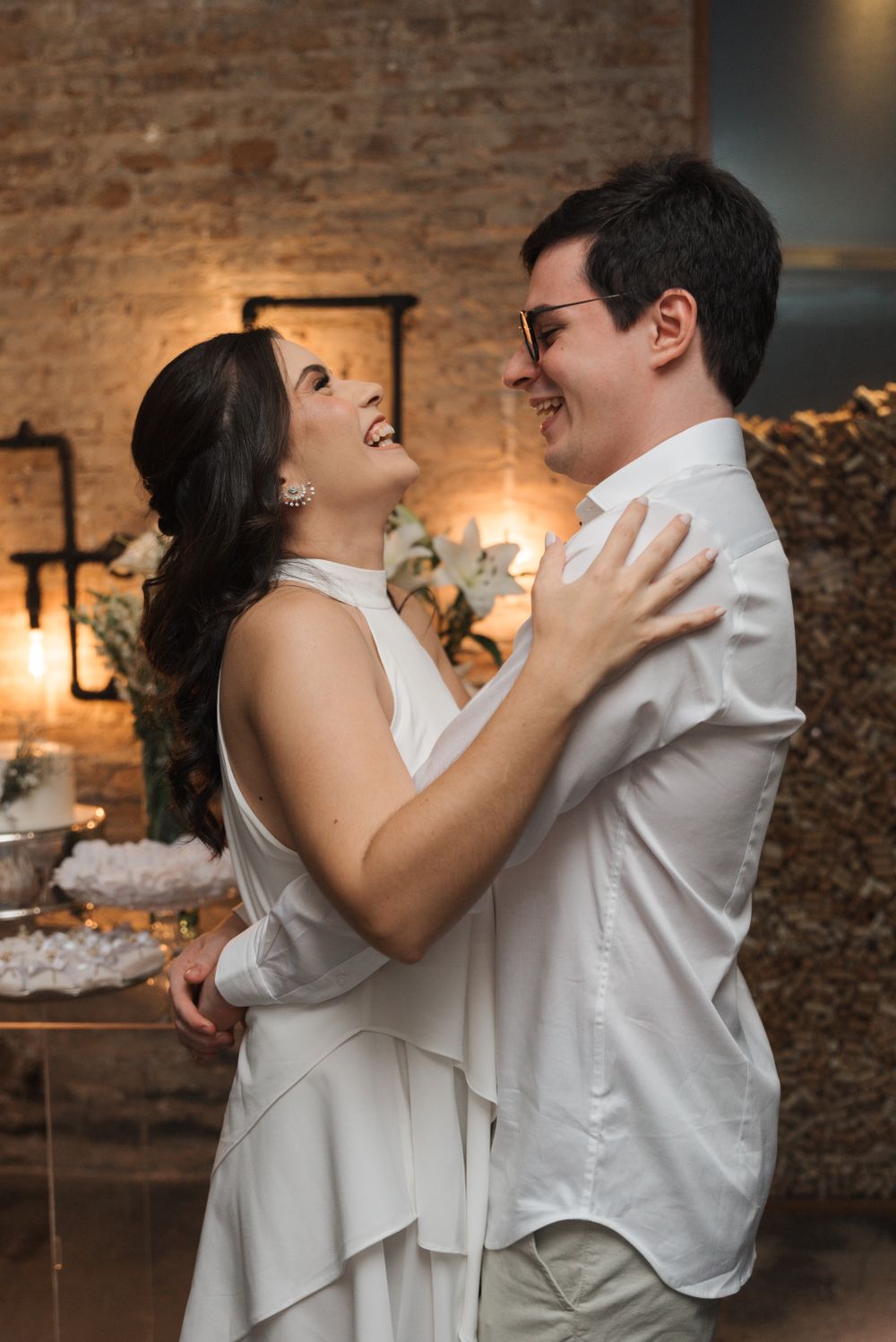 A joyful couple sharing a laugh while embracing in formal white attire against a rustic brick wall.
