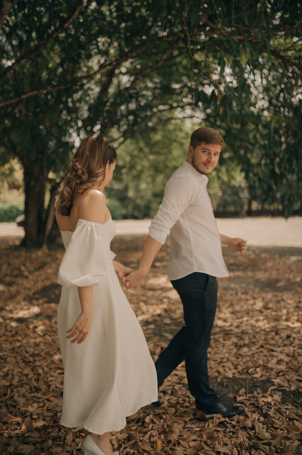 A couple in coordinated white and navy outfits walking through fallen leaves under trees.