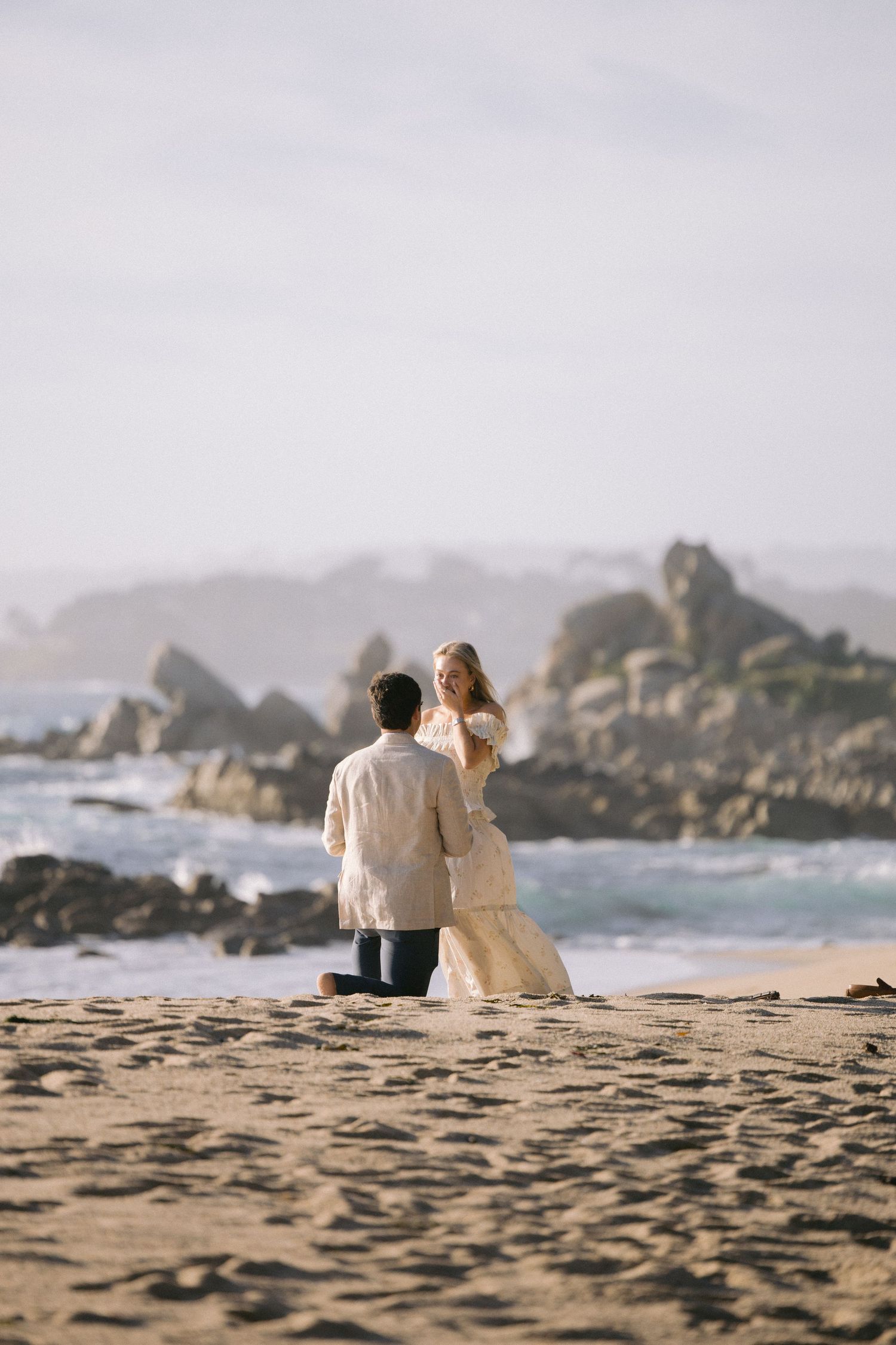 Two people sitting together on a sandy beach with rocky coastal cliffs and ocean waves in the background.