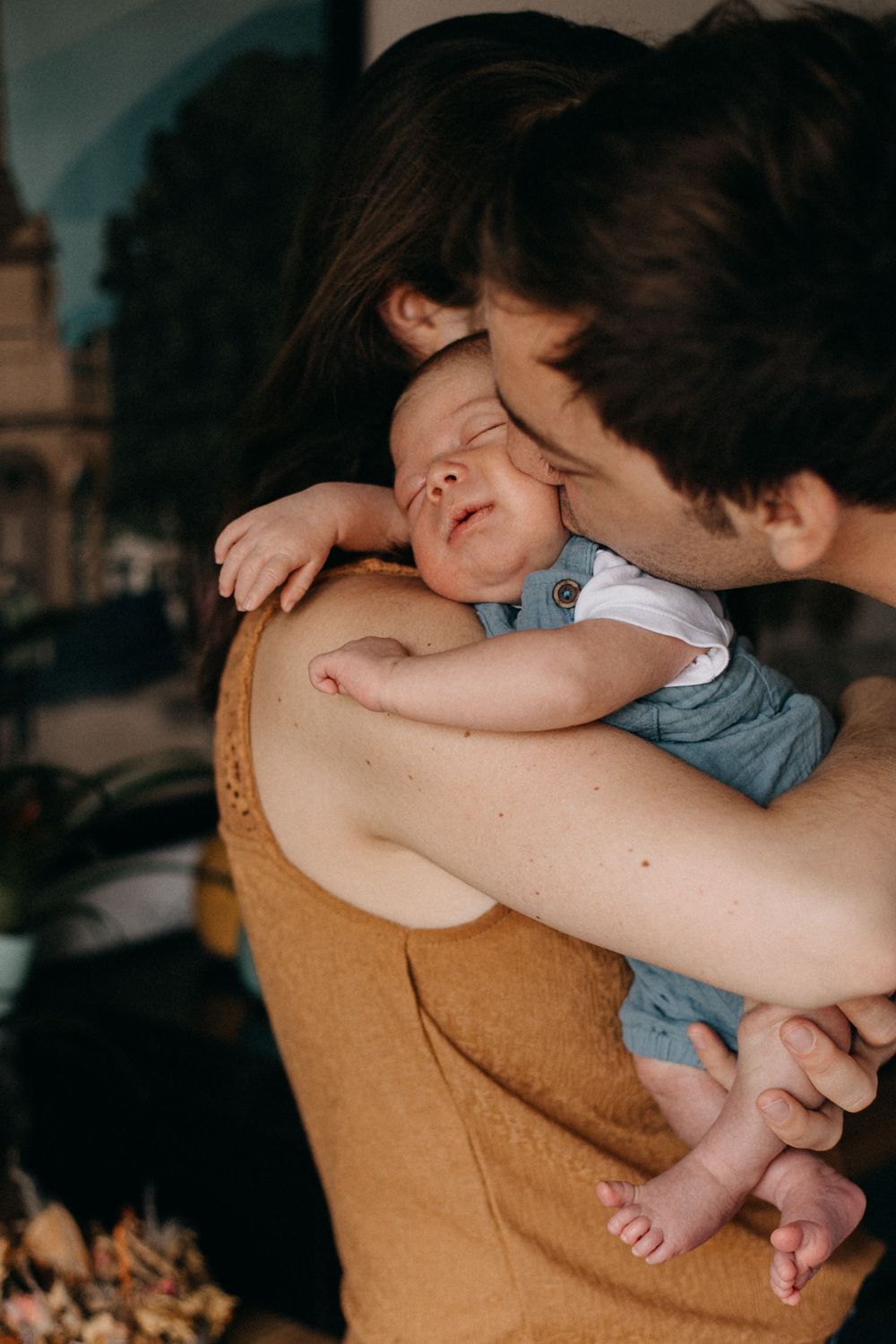 Parent holds and kisses sleeping baby in casual clothing against warm toned background.