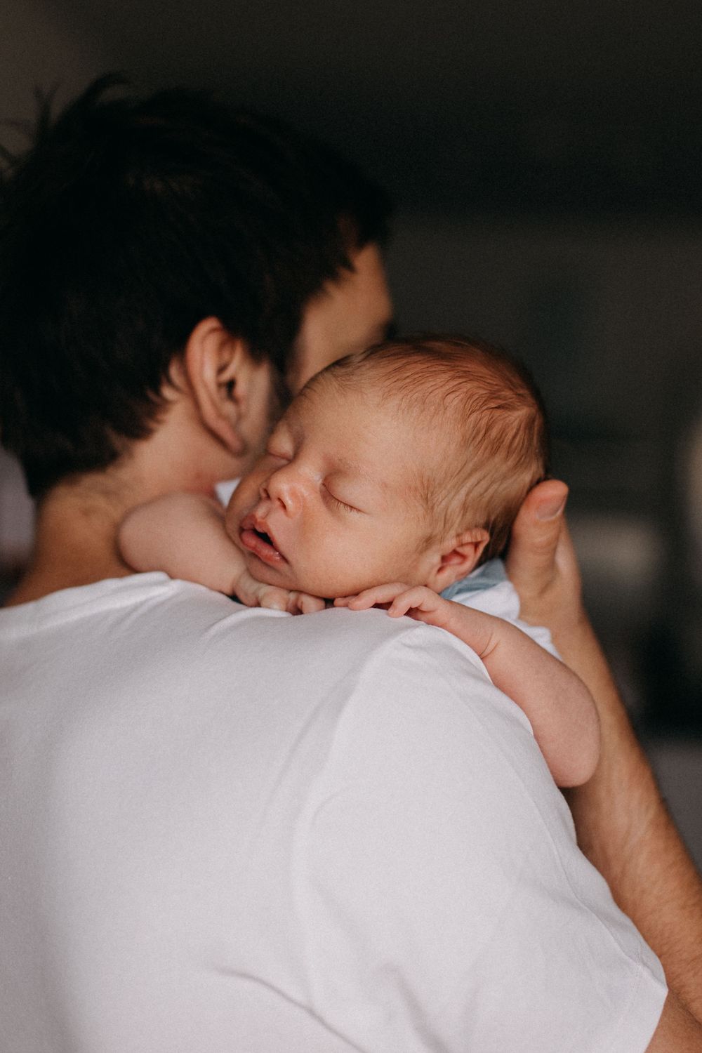 A tender moment between parent and sleeping newborn baby resting on shoulder in soft lighting.
