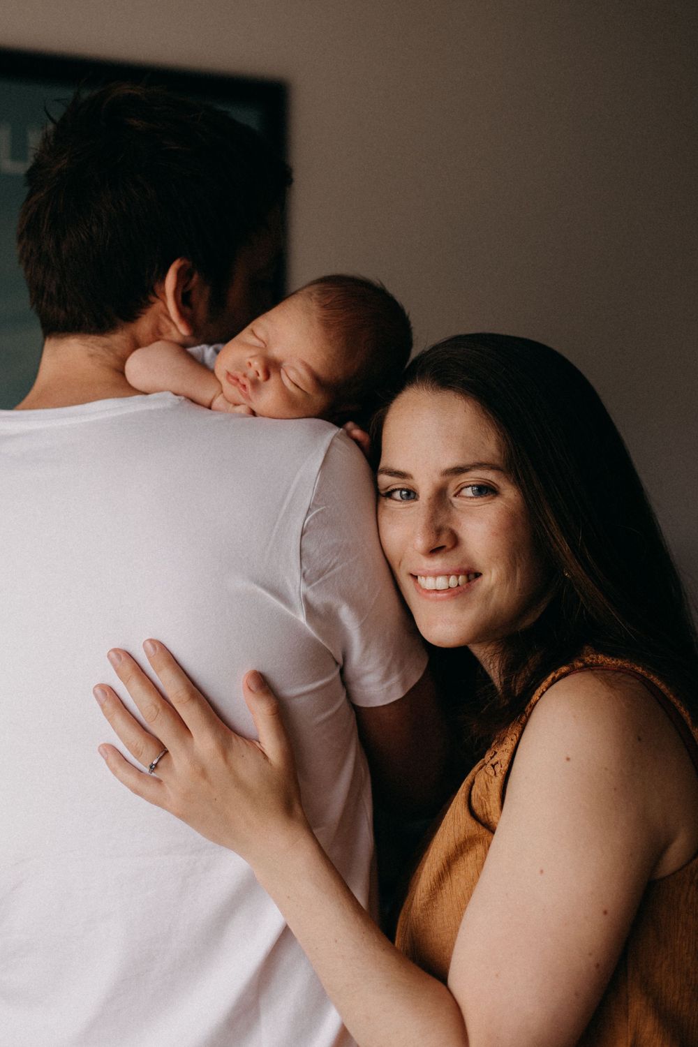 Parents share tender moment while holding sleeping newborn baby on shoulder.