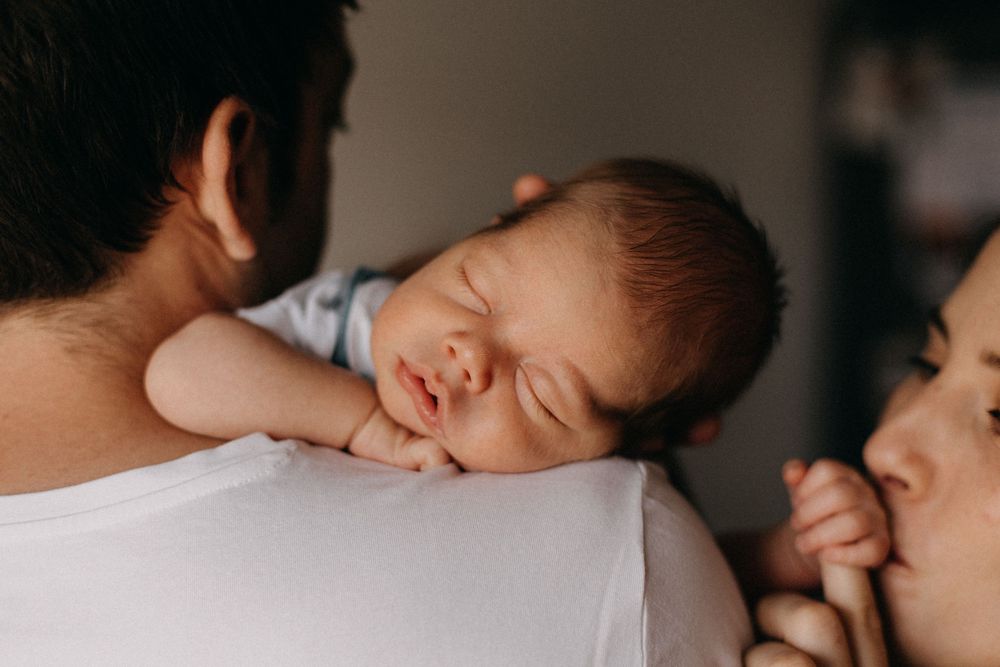 A sleeping newborn baby rests peacefully on a parent's shoulder in a tender family moment.