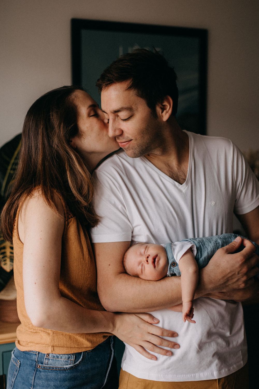 Intimate family moment as parents share kiss while holding sleeping newborn baby.