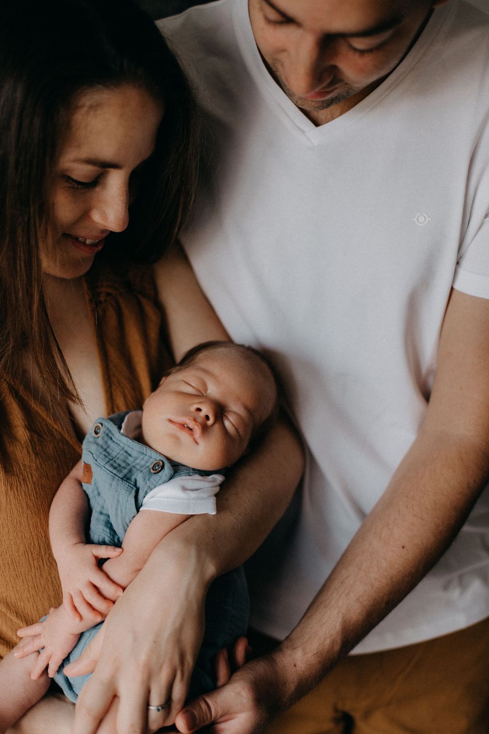 A sleeping baby in denim overalls nestles peacefully between two adults in a warm-toned photograph.