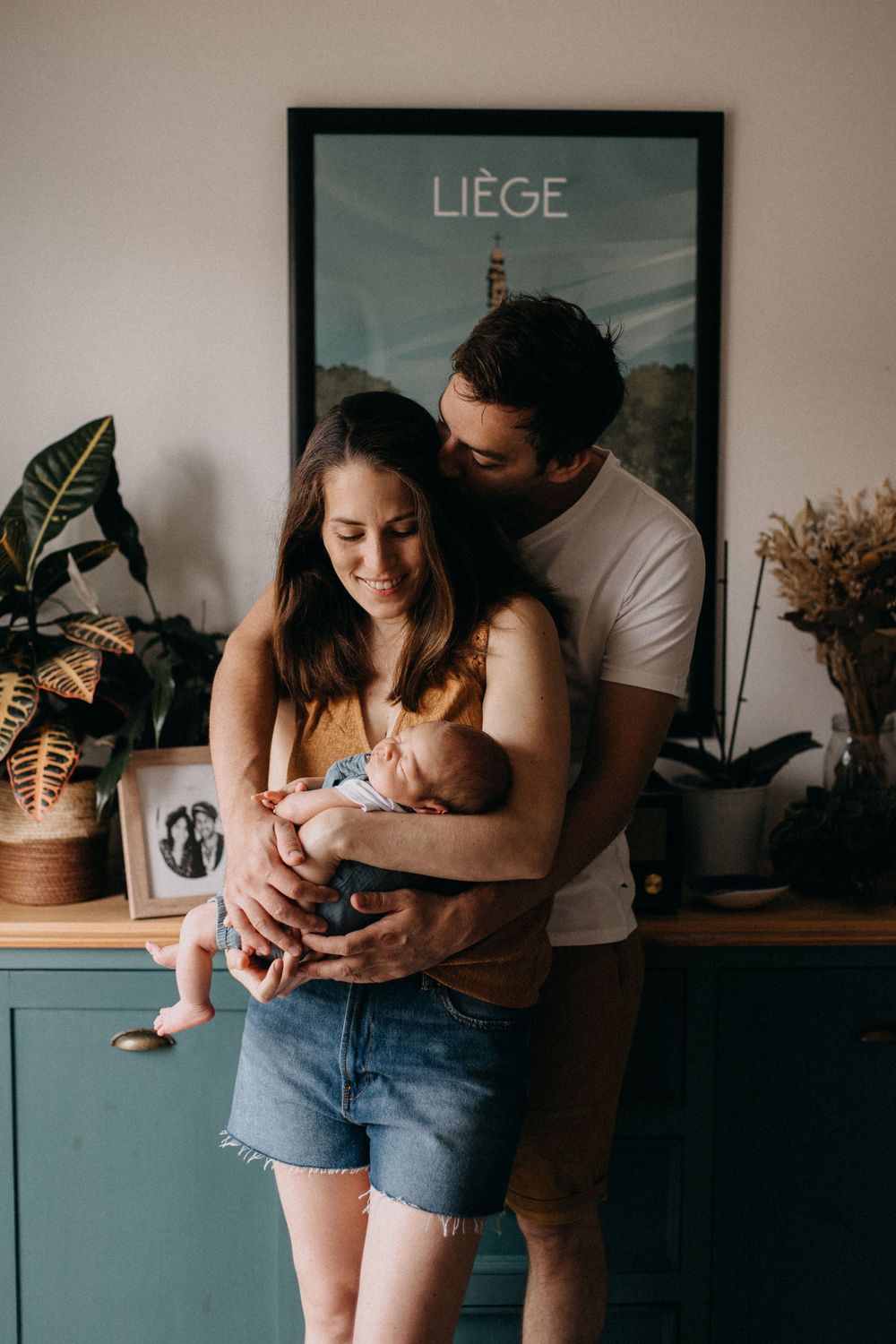 Intimate family moment in a cozy home interior with a newborn baby and houseplants against a Liège poster.