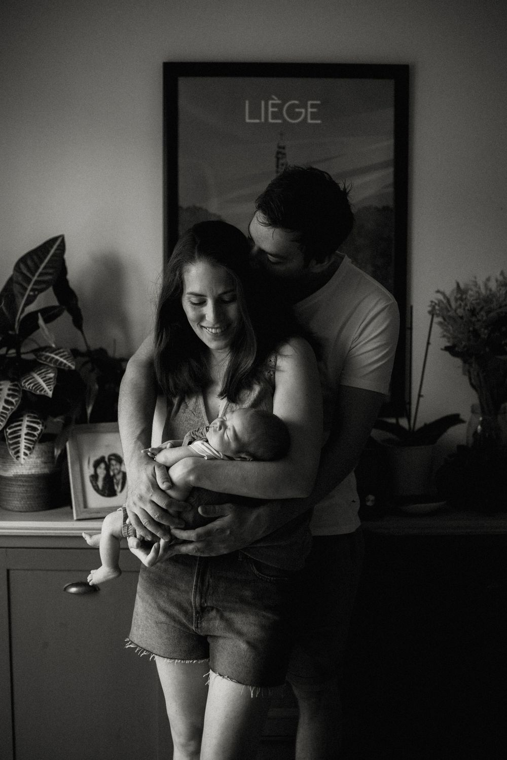 A tender family moment captured in black and white with a Liege poster and plants in the background.