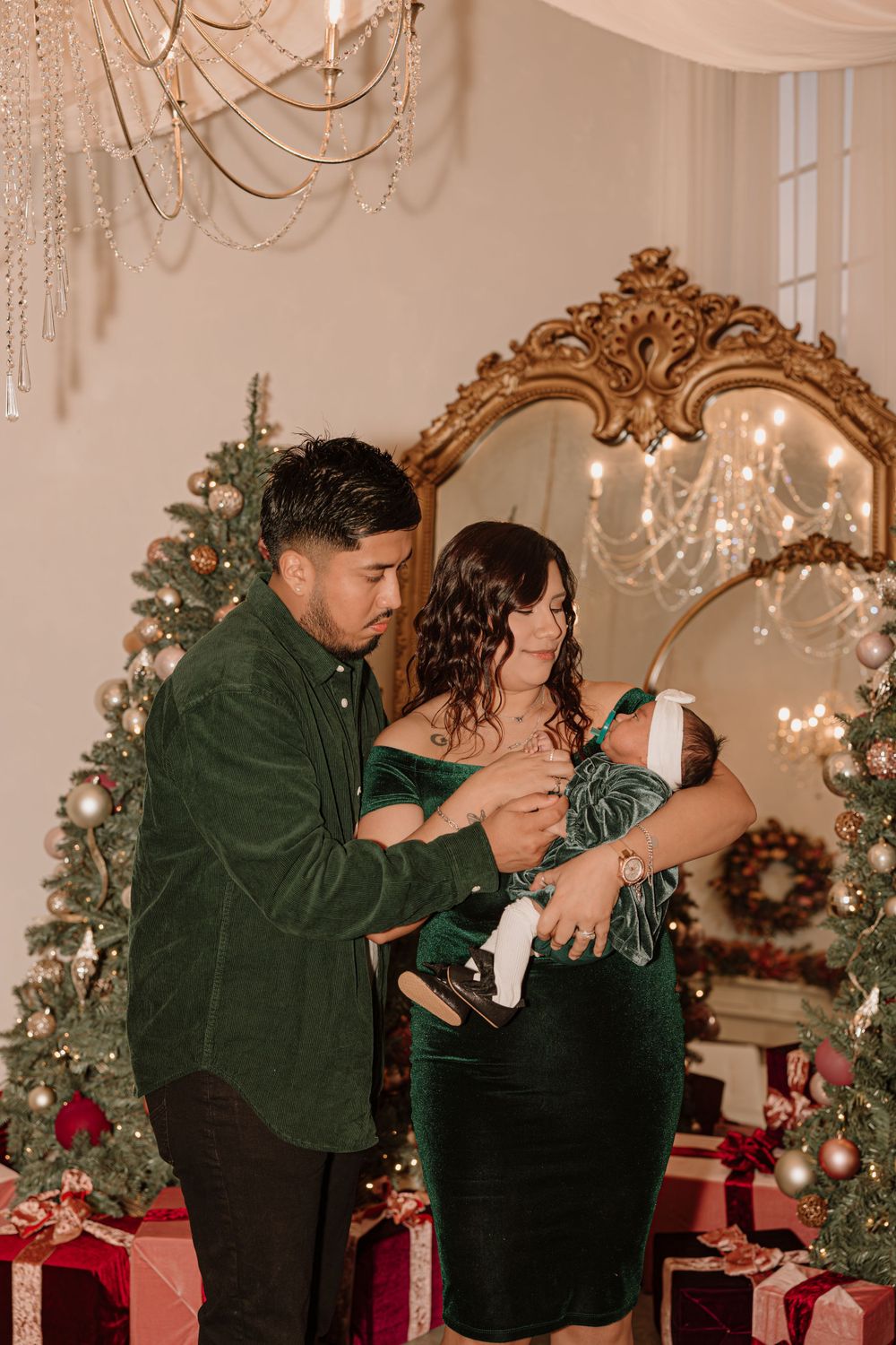 A couple in formal attire tends to their baby in front of decorated Christmas trees and a gold mirror.