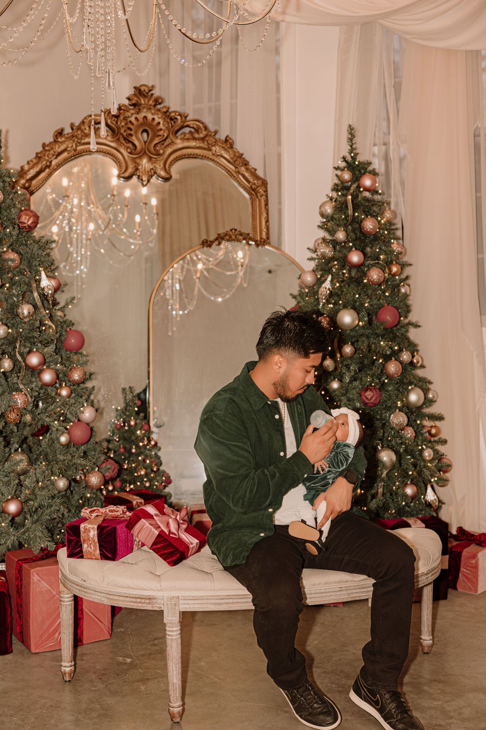 An elegant holiday scene with Christmas trees and a person holding an infant on an ornate bench.