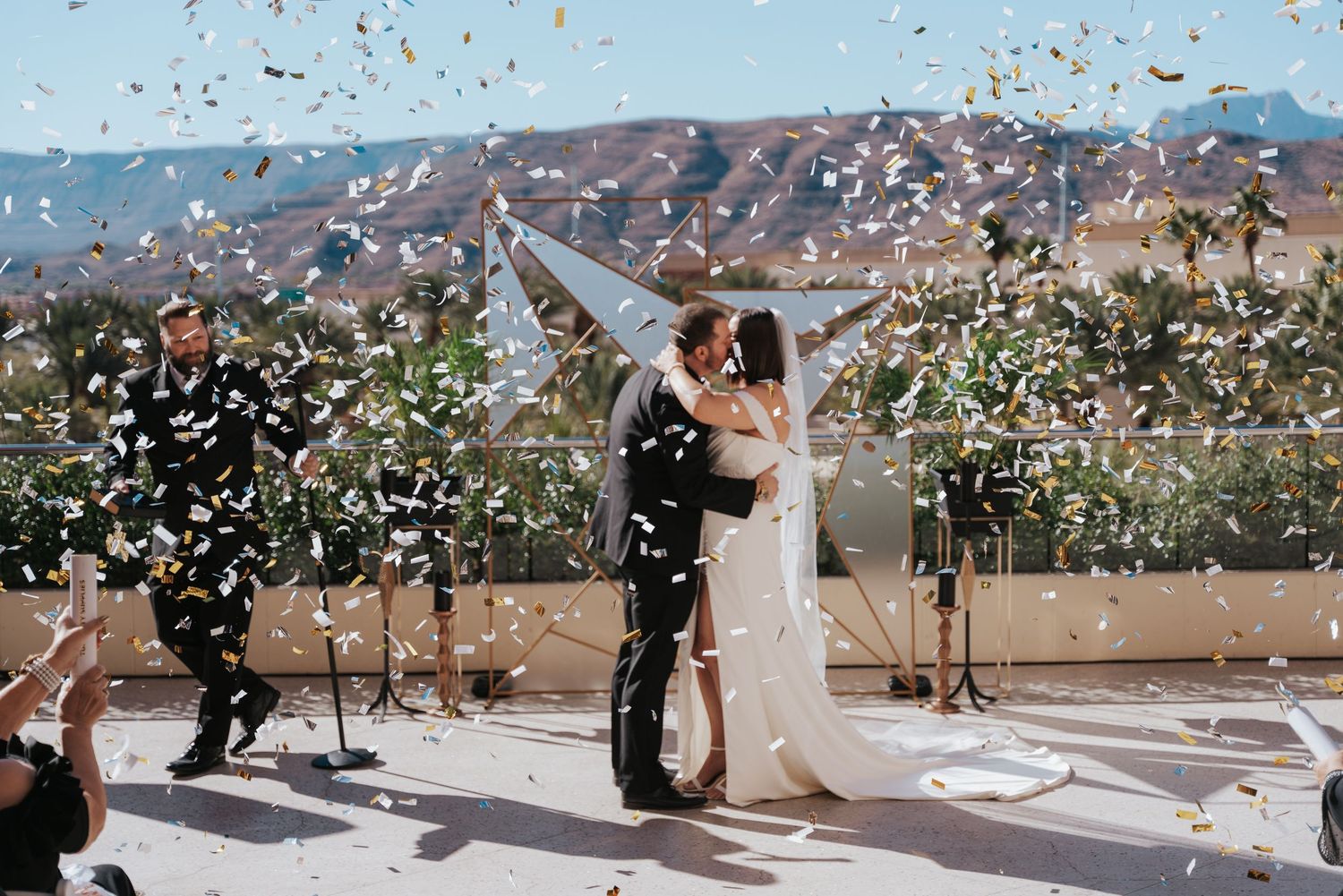 A couple embraces during their wedding ceremony with confetti floating through the air against scenic mountain views.