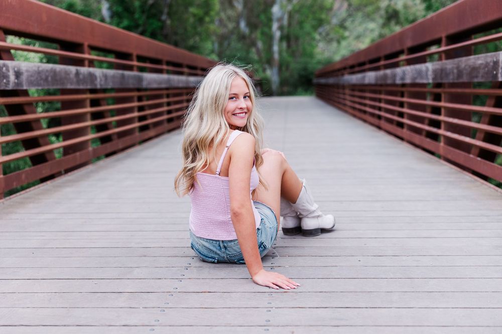 A person sits casually on a wooden bridge surrounded by trees on a bright summer day.
