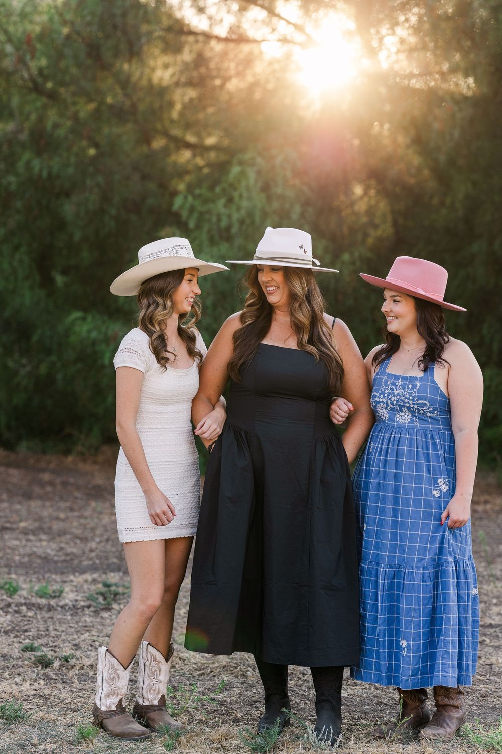 Three friends in western style dresses and hats standing together outdoors at sunset with golden sunlight in background.