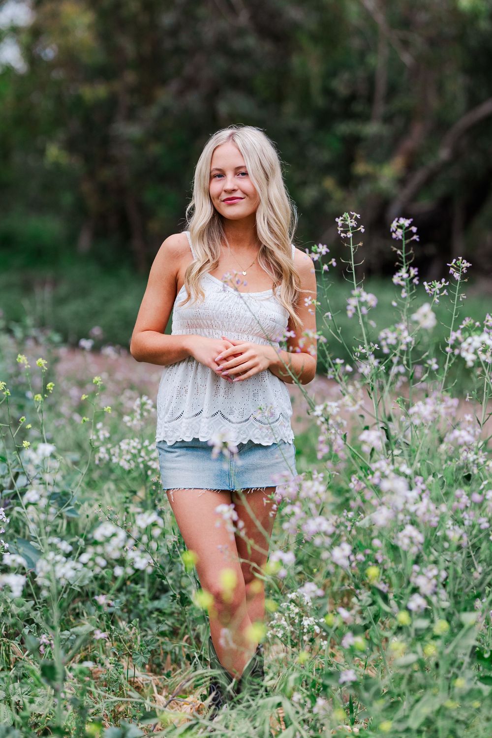 Woman wearing white lace top and denim shorts poses in a meadow of wildflowers on a sunny summer day.
