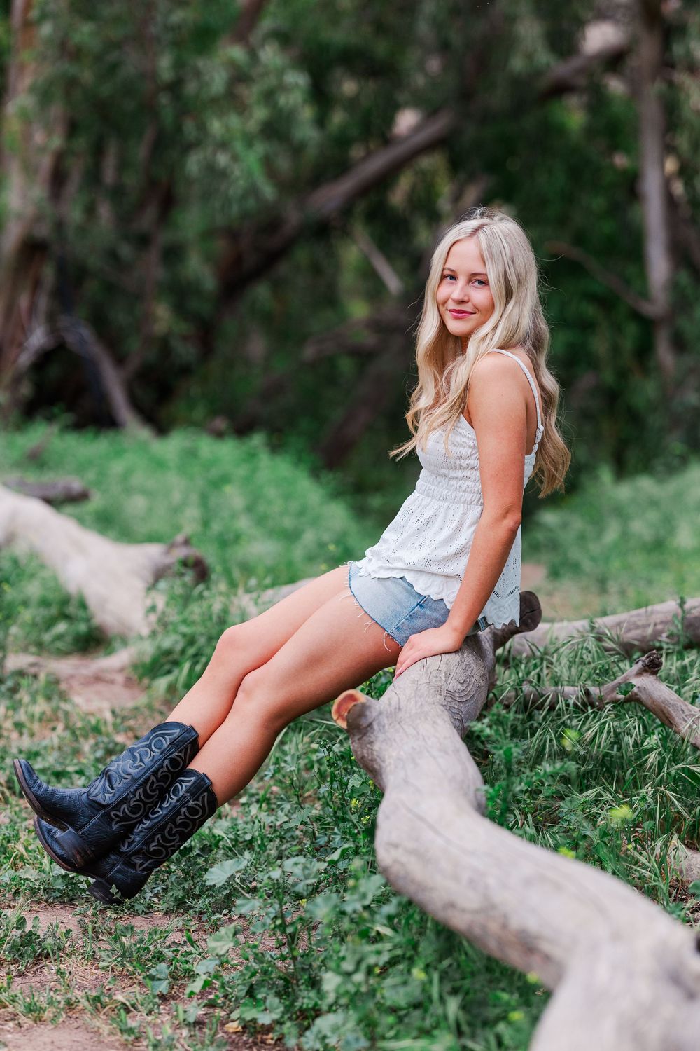 A person in a white top and denim shorts sits on a fallen tree trunk wearing black combat boots in a woodland setting.