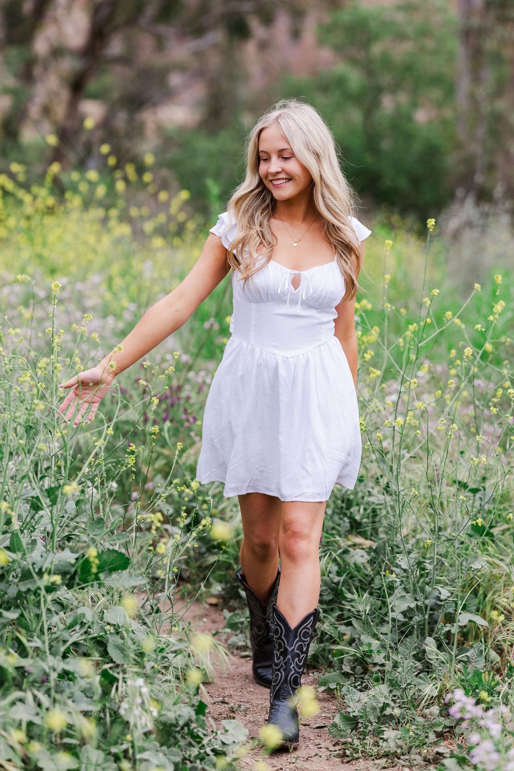 A woman in a white sundress and cowboy boots walks through a meadow of yellow wildflowers on a sunny day.