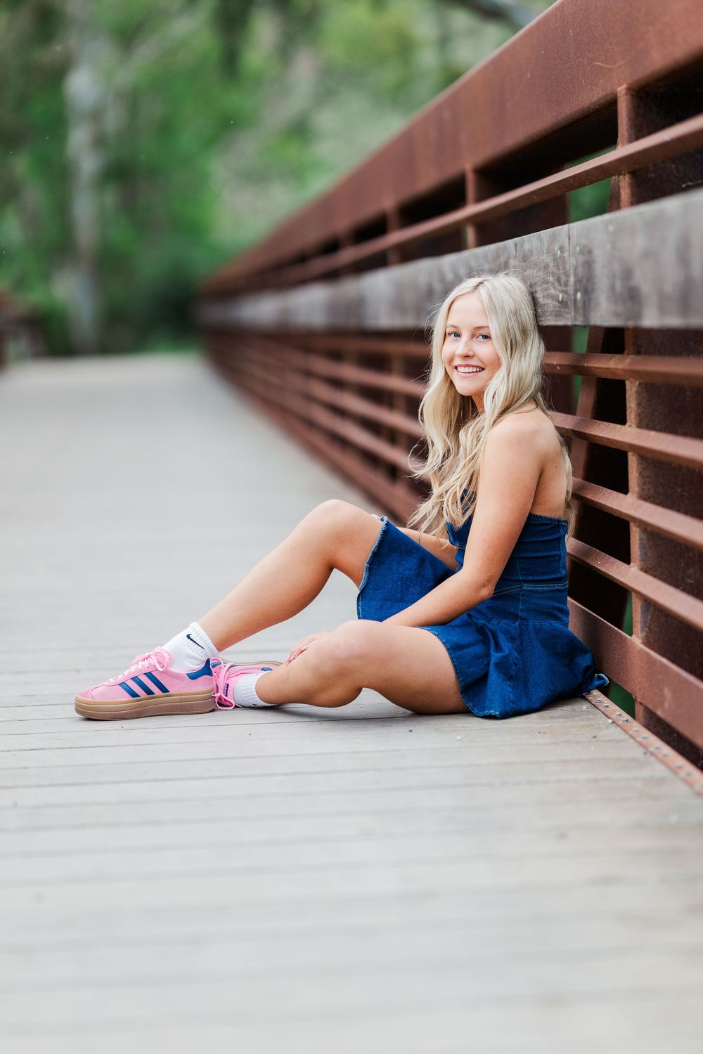 A person in a navy blue dress poses casually on a wooden bridge with pink athletic shoes on a sunny day.