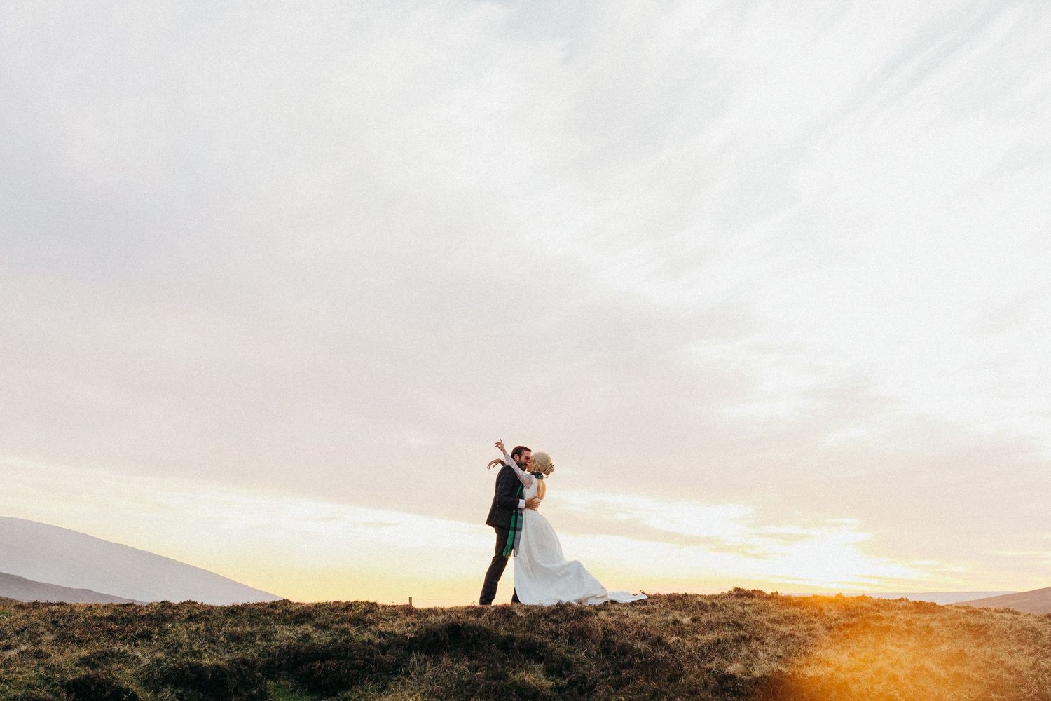 A couple embraces on a hilltop during a golden sunset with mountains in the background.