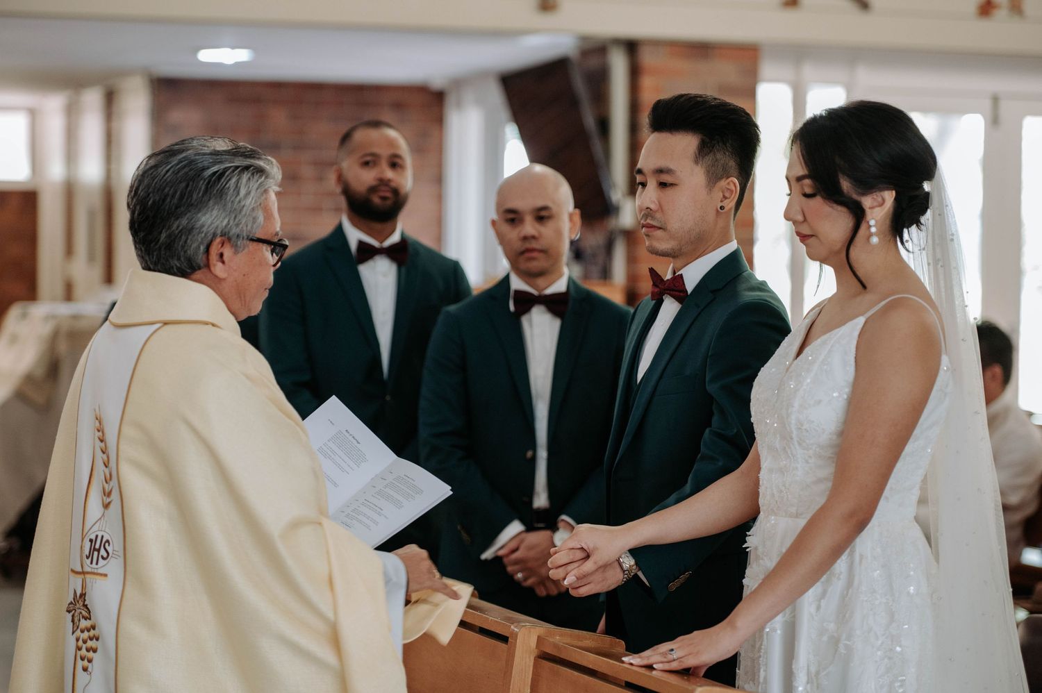 A couple holds hands during their wedding ceremony as they listen to the officiating priest in a church setting.