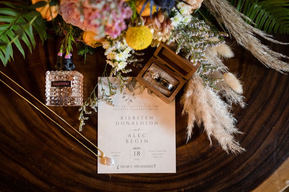 Wedding invitation suite with perfume bottles and dried flowers arranged on dark wooden surface with elegant styling.