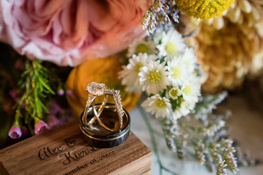 Wedding rings rest on wooden surface beside pink roses and white daisies in floral arrangement.