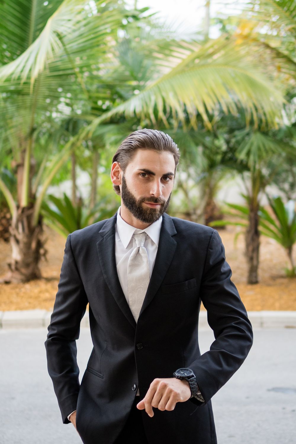 Formal suited man checks time while standing near tropical palm trees in outdoor setting.