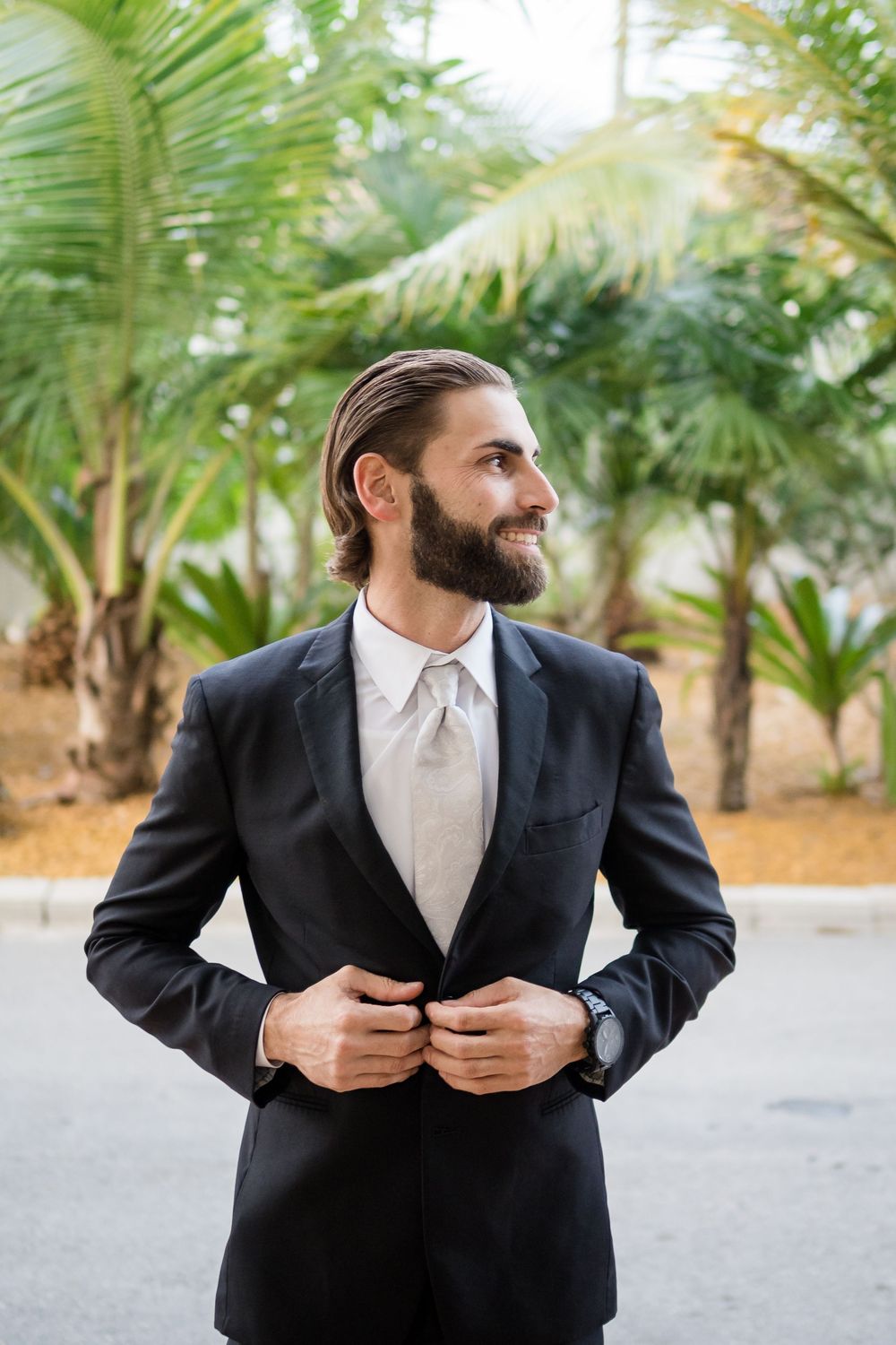 Bearded groom in black suit and light tie poses outdoors with palm trees in background.
