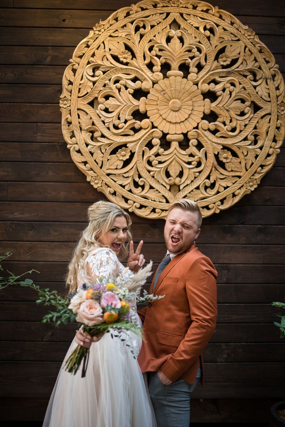 A couple poses playfully in front of a large decorative wooden mandala wall art at their wedding.